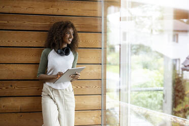 Smiling woman with digital tablet looking through window while standing at home - PESF02222