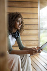 Junge Frau mit Kopfhörer, die zu Hause sitzend ein digitales Tablet benutzt - PESF02224