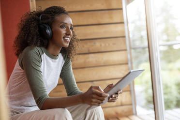 Lächelnde Frau mit Kopfhörern, die ein digitales Tablet benutzt, während sie zu Hause sitzt - PESF02226