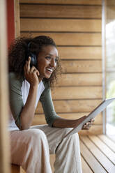 Lächelnde Frau, die über Kopfhörer spricht, während sie zu Hause ein digitales Tablet benutzt - PESF02228