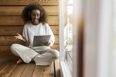 Junge Frau mit Kopfhörern, die ein Videogespräch über ein digitales Tablet führt, während sie zu Hause am Fenster sitzt - PESF02234