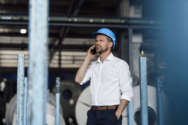 Männlicher Unternehmer, der in einer Fabrik mit der Hand in der Tasche steht und mit einem Smartphone spricht - GUSF04721