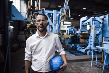 Male entrepreneur wearing protective eyewear while holding helmet standing in factory - GUSF04729