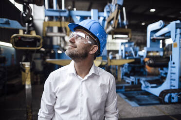 Lächelnder männlicher Unternehmer, der mit einer Schutzbrille in einer Fabrik nach oben schaut - GUSF04733