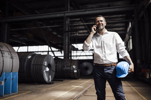 Lächelnder Geschäftsmann, der mit seinem Smartphone spricht, während er in der Industrie steht - GUSF04750