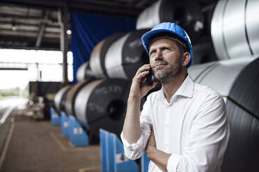 Male owner talking on smart phone looking up while standing against steel rolls in industry - GUSF04765