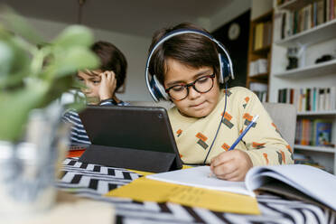 Junge, der mit einem männlichen Freund beim E-Learning zu Hause sitzt und über ein digitales Tablet Notizen schreibt - VABF04031