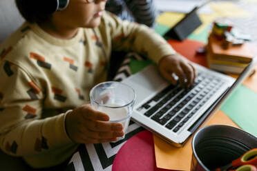 Boy holding glass of milk while sitting with laptop at home - VABF04045