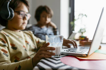 Junge hält ein Glas Milch in der Hand, während er zu Hause einen Laptop benutzt - VABF04046