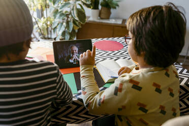 Elementary age boy pointing while e-learning through video call sitting with male friend in living room - VABF04058