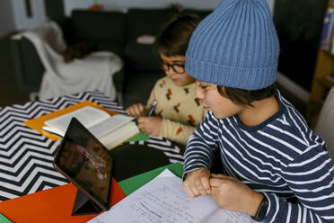 Boy talking to teacher during e-Learning class while male friend studying beside in living room - VABF04059