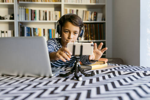 Junge, der sein Smartphone auf einem Stativ neben einem Laptop am Tisch einstellt - VABF04065