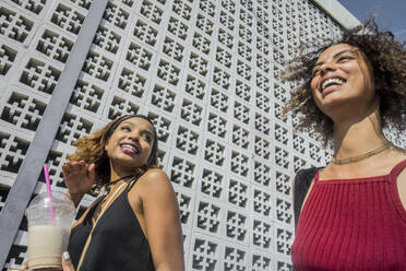 Cheerful young female friends walking by wall on sunny day - AJOF00616