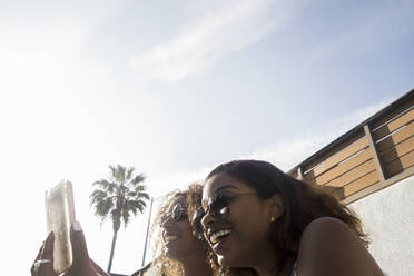 Glückliche junge Frauen teilen Erinnerungen auf dem Smartphone gegen den Himmel an einem sonnigen Tag - AJOF00621