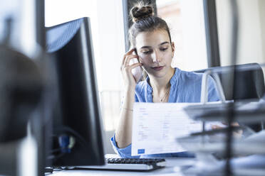 Junge Geschäftsfrau hält Papier, während sie im Büro mit dem Handy telefoniert - UUF22076