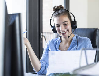 Businesswoman with paper talking through headphones while sitting at office - UUF22081