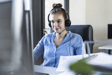 Junge Geschäftsfrau mit Kopfhörern, die am Computer arbeitet, während sie im Büro sitzt - UUF22082