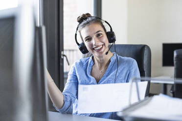 Glückliche Geschäftsfrau mit Kopfhörern, die im Büro sitzt und arbeitet - UUF22085