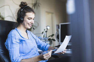 Young businesswoman wearing headphones holding paper while sitting on chair at office - UUF22087