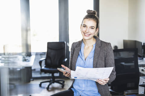 Lächelnde Geschäftsfrau mit Papier, die ein Mobiltelefon benutzt, während sie im Büro steht, lizenzfreies Stockfoto