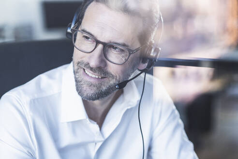 Lächelnder Geschäftsmann mit Kopfhörern, der wegschaut, während er im Büro sitzt - UUF22096