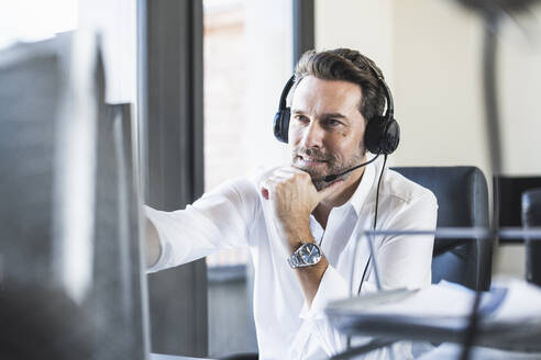 Geschäftsmann mit Kopfhörern, der mit der Hand am Kinn sitzt, während er am Computer im Büro arbeitet - UUF22098
