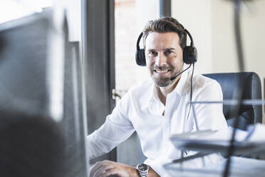 Smiling businessman wearing headphones sitting at office - UUF22099