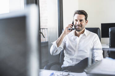 Mature businessman talking on mobile phone while sitting on chair at office - UUF22100