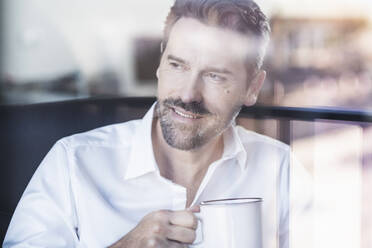 Smiling businessman drinking coffee while sitting at office - UUF22104