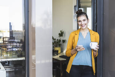 Businesswoman with coffee cup using mobile phone while standing at door in office - UUF22110