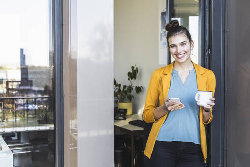 Geschäftsfrau mit Kaffeetasse, die ein Mobiltelefon benutzt, während sie an der Tür im Büro steht - UUF22110