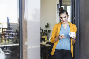 Businesswoman with coffee cup smiling while using mobile phone standing at office - UUF22111