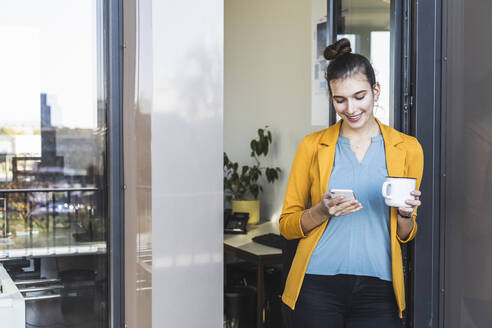 Geschäftsfrau mit Kaffeetasse, die lächelt, während sie im Büro steht und ein Mobiltelefon benutzt - UUF22111