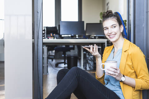 Geschäftsfrau mit Kaffeetasse, die mit ihrem Handy telefoniert, während sie am Eingang eines Büros sitzt, lizenzfreies Stockfoto