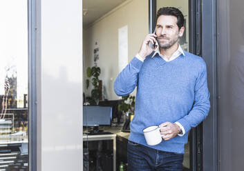 Älterer Geschäftsmann mit Kaffeetasse, der mit seinem Handy telefoniert, während er am Büroeingang steht - UUF22115