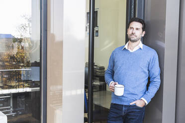 Businessman holding coffee cup while leaning on wall with hand in pocket at office entrance - UUF22116