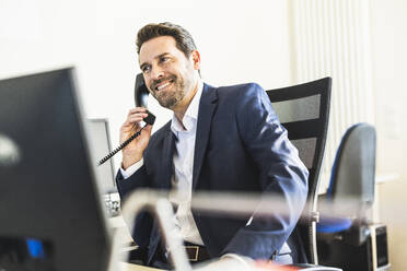 Lächelnder Geschäftsmann, der auf einem Stuhl sitzend im Büro telefoniert - UUF22124