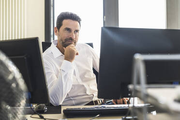 Geschäftsmann mit Hand am Kinn, der wegschaut, während er im Büro sitzt - UUF22128