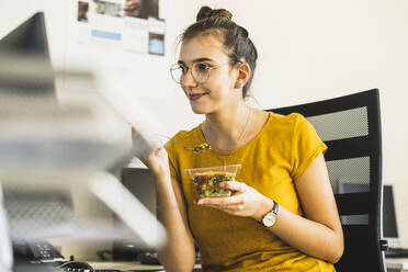 Lächelnde Geschäftsfrau, die Essen isst, während sie im Büro am Computer sitzt - UUF22134