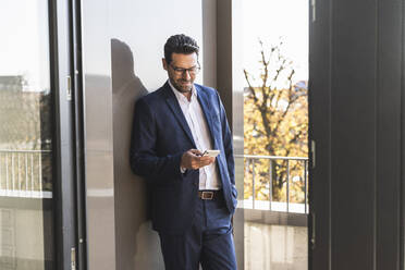 Male entrepreneur using mobile phone while standing with hand in pocket by balcony at office - UUF22142