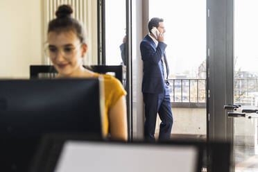 Colleague working on computer while sitting in office with businessman standing in background by balcony - UUF22145