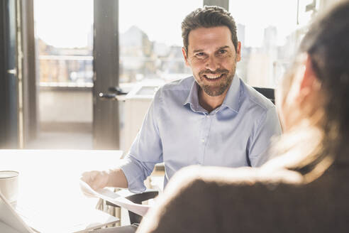 Lächelnder Geschäftsmann bei der Arbeit mit einem Kollegen, während er im Büro sitzt - UUF22167
