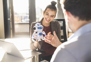 Junge Geschäftsfrau zeigt einem Geschäftsmann ein Roboterspielzeug, während sie im Büro sitzt - UUF22177