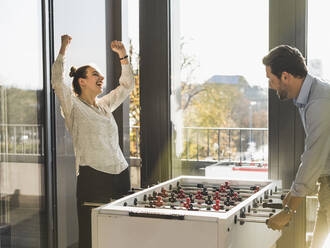 Glückliche Geschäftsfrau zeigt gewinnende Geste beim Kickerspiel mit Geschäftsmann im Büro - UUF22185