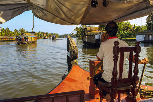 Der Kapitän telefoniert und steuert das Hausboot durch die Backwaters auf einer beliebten Backwater-Kreuzfahrt, Alappuzha (Alleppey), Kerala, Indien, Asien - RHPLF17994