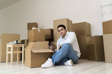 Junger Mann, der ein Mobiltelefon benutzt, während er gegen Kartons in einem neuen Haus sitzt - GIOF09698