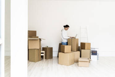 Young man working on laptop while standing amidst cardboard boxes in new apartment - GIOF09701