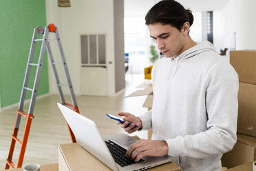 Junger Mann bei der Arbeit am Laptop und der Benutzung eines Mobiltelefons während des Umzugs in ein neues Haus - GIOF09703