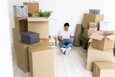Man working on laptop surrounded by boxes while sitting in new apartment - GIOF09705