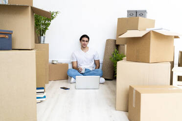 Young man drinking coffee while sitting on floor surrounded by boxes in new apartment - GIOF09706